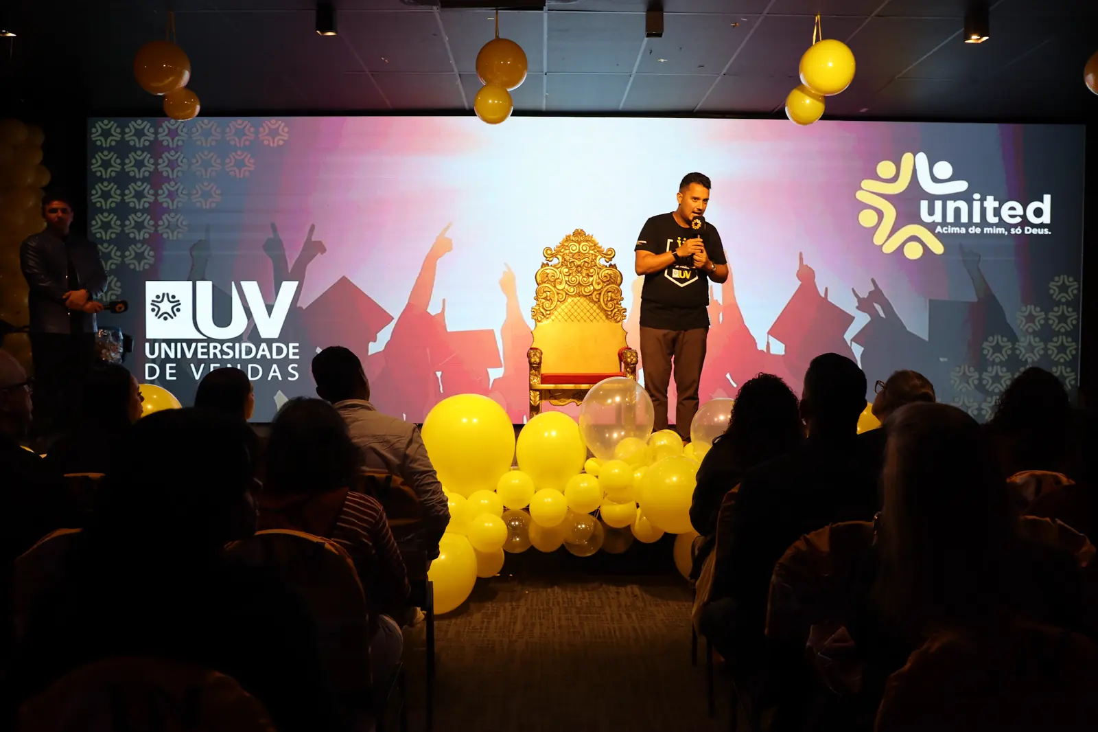 Palco do Centro de Convenções com os logos da Universidade de Vendas e da United Class, balões amarelos e a plateia em silhueta